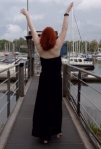 Harbour setting, a woman with red hair in the Center of the picture. Her back is turned, she raises her arms over her head. Her long black dress floats gently in the wind